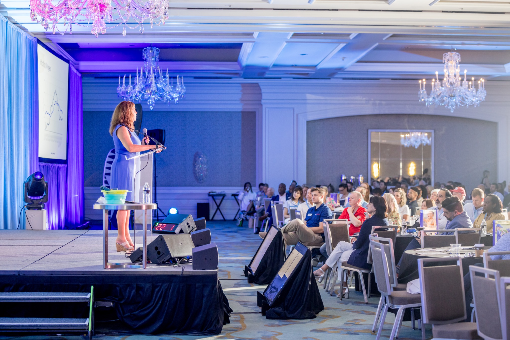Presenter in blue on a lit stage speaking to a seated conference audience, photographed by Ti Adoro Studios